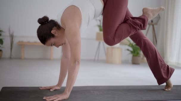 Young woman in comfortable sportswear, leggings and top practising yoga in modern studio on mat with white walls and plants, sporty Latina girl doing Dhanurasana, bow pose, workout and wellness. - Footage, Video