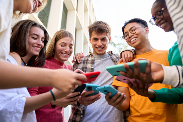 Group of multiracial generation z young smile cheerful people playing funny videos game on a smart phone they holding in their hands. Friends having fun together using social on mobile cell outdoor - Photo, Image