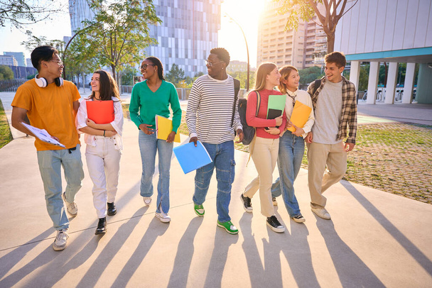 Boldog nemzetközi diákok sétálgatnak az egyetem területén. Fiatal multikulturális barátok mosolyognak és nézik egymást hátizsákkal és munkafüzetekkel a szabadban - Fotó, kép