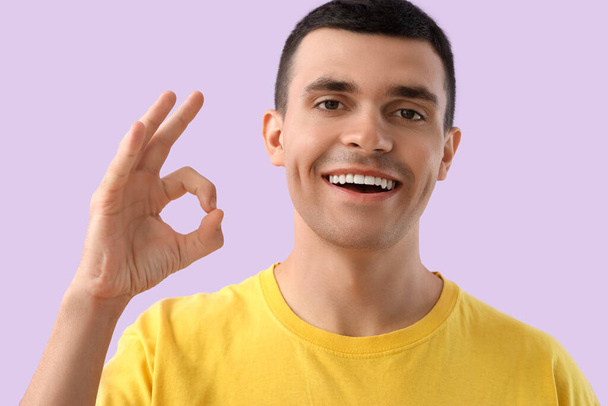 Красивый молодой человек со здоровыми зубами показывает ОК жест на фиолетовом фоне. Концепция стоматологической помощи - Фото, изображение