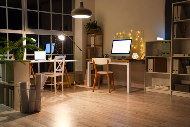 Interior de la oficina con escritorios y lámparas brillantes por la noche - Foto, Imagen