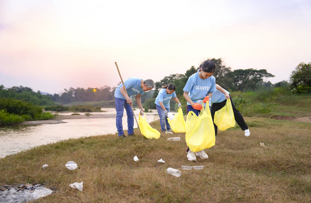 grupa wolontariuszy w niebieskim t-shircie zbierająca śmieci i sortująca odpady z tworzyw sztucznych, dobrowolna grupa rodzinna sprzątająca brudne miejsce publiczne pełne garbusów nad rzeką w wieczornym słońcu - Zdjęcie, obraz