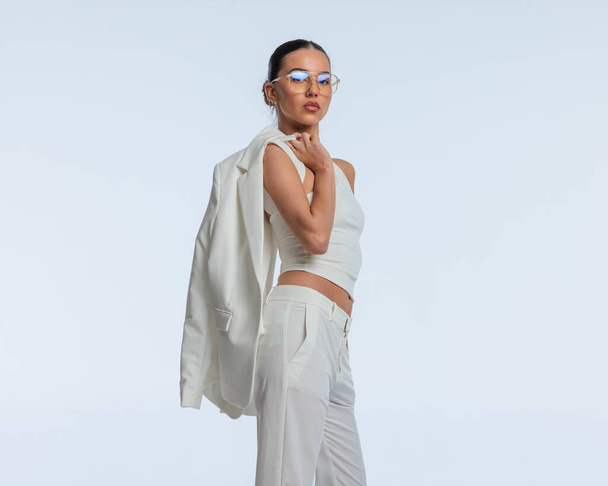 крутая элегантная бизнесвумен в очках с пиджаком через плечо, смотрящая вперед и сидящая перед серым фоном - Фото, изображение