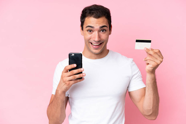 Joven hombre caucásico aislado sobre fondo rosa comprando con el móvil y sosteniendo una tarjeta de crédito con expresión sorprendida - Foto, imagen