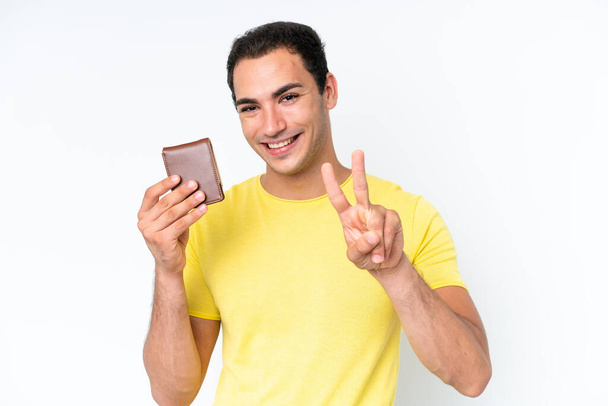 Mladý běloch drží peněženku izolovanou na bílém pozadí, usmívá se a ukazuje znamení vítězství - Fotografie, Obrázek