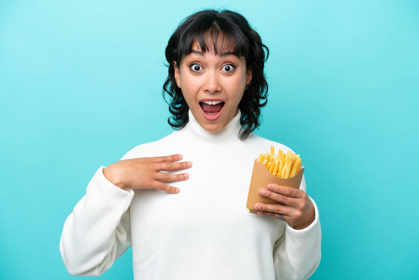 Молодая аргентинская женщина с жареными чипсами на синем фоне удивлена и шокирована, глядя направо - Фото, изображение