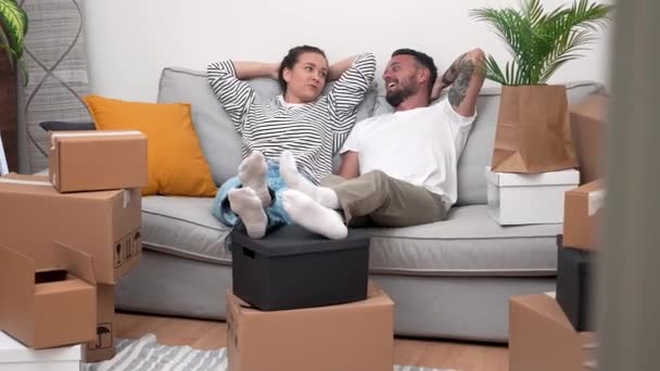 κουρασμένο αλλά ενθουσιασμένο ζευγάρι γέρνει πίσω στον καναπέ, που περιβάλλεται από κινούμενα κουτιά χαρτοκιβωτίων, συζήτηση για διακόσμηση σπιτιού και ανακαινίσεις στο νέο τους διαμέρισμα - Πλάνα, βίντεο
