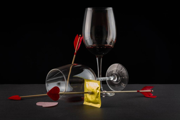ένα προφυλακτικό τρυπημένο από βέλος του έρωτα με ποτήρια κρασιού σε μαύρο φόντο - Φωτογραφία, εικόνα