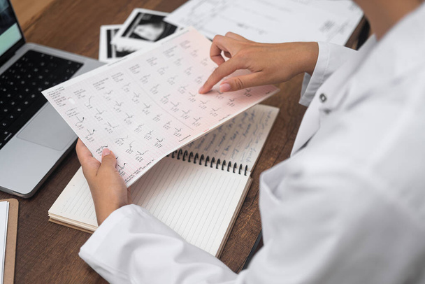 Молодий лікар читає документи з результатами тестів на кардіограму пацієнта жінка-кардіолог вивчає документи, що сидять за столом з комп'ютером і видом збоку стетоскопа - Фото, зображення