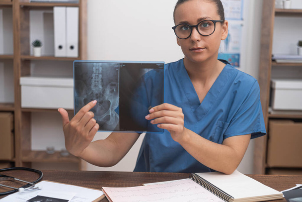 Mulher traumatologista aponta dedo para imagem de raio-X conversando com paciente on-line olhando para câmera médico consulta visitante remotamente tratamento na clínica moderna - Foto, Imagem
