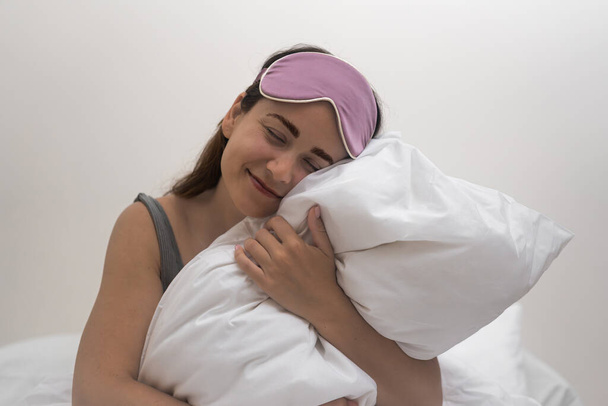 Nel santuario della sua camera da letto, una signora affascinante con una maschera del sonno sorride, custodendo un soffice cuscino bianco al mattino..  - Foto, immagini