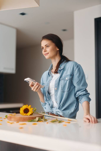 Νεαρή καυκάσια γυναίκα σε μια σύγχρονη κουζίνα, κρατώντας ένα Smartphone και απολαμβάνοντας ένα υγιεινό πρωινό - Φωτογραφία, εικόνα