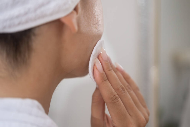 A serene close-up: Towel-wrapped woman gently applies cleansing product to her face with a cotton pad, studying her reflection.  - Photo, Image