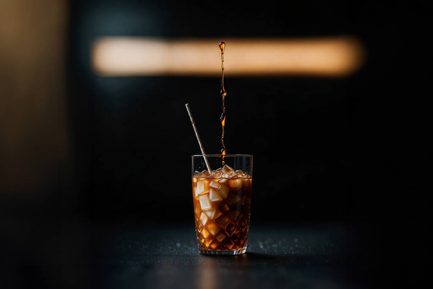 an iced coffee glass against a dark background. The rich brown coffee, seen through frosted glass, creates a visually striking composition. Ice cubes float in the coffee, adding a refreshing touch. The dark, almost black background enhances the visua - Photo, Image