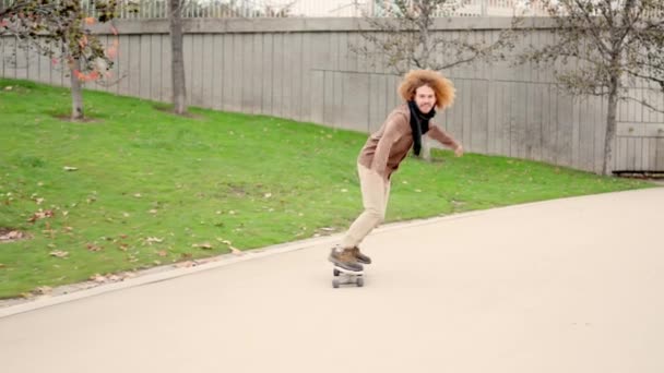 Slow motion video alternatywnego młodego człowieka z kręconymi włosami na deskorolce w n miejskim parku - Materiał filmowy, wideo
