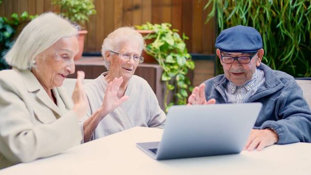 Trzech seniorów korzystających z laptopa i machających podczas rozmowy wideo na zewnątrz w domu opieki - Zdjęcie, obraz