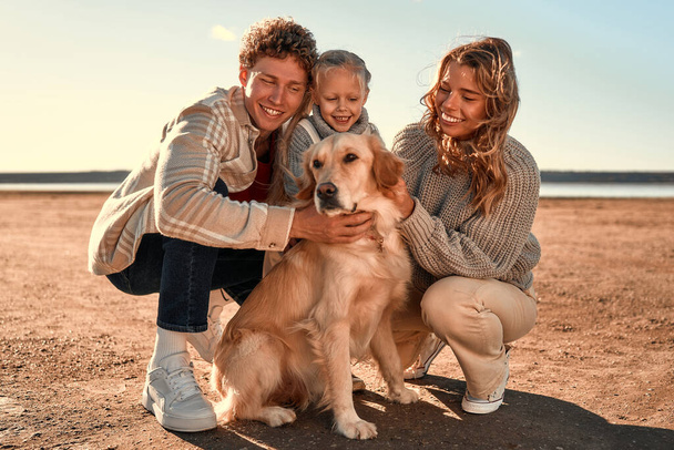Um jovem casal de pais com uma pequena filha fofa e seu cão retriever amado andando e relaxando em uma praia de areia em tempo fresco em seu fim de semana. Família viajando com animal de estimação. - Foto, Imagem