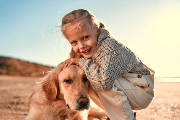 Cute little girl child having fun and playing with her golden retriever dog outside. Girl with a dog on a sandy beach. - Photo, Image