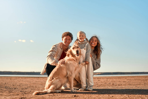 Una giovane coppia di genitori con una piccola figlia carina e il loro amato cane retriever passeggiando e rilassandosi su una spiaggia di sabbia con il tempo fresco nel loro fine settimana. Famiglia in viaggio con animale domestico. - Foto, immagini