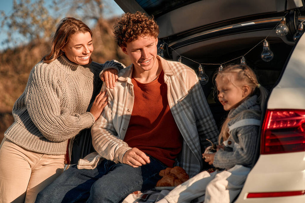 Молода щаслива сім'я на пікніку зі своєю улюбленою маленькою донькою сидить в задній частині машини на ковдрі у вихідні, розважаючись разом. - Фото, зображення