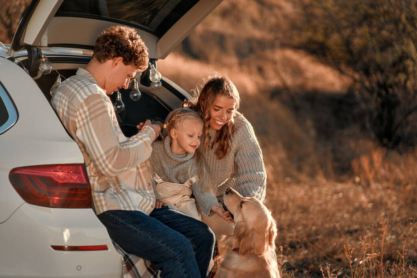 Una joven familia feliz en un picnic con su querida hija y su perro sentados en la parte trasera de un coche en una manta en un fin de semana divirtiéndose juntos. - Foto, Imagen