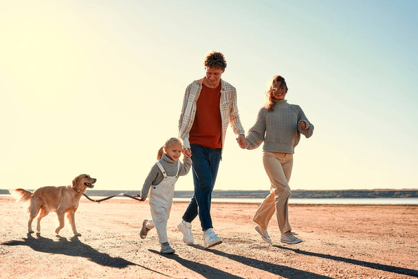 Una joven pareja de padres con una pequeña hija linda y su querido perro recuperador paseando y relajándose en una playa de arena en tiempo fresco en su fin de semana. Familia viajando con mascota. - Foto, imagen