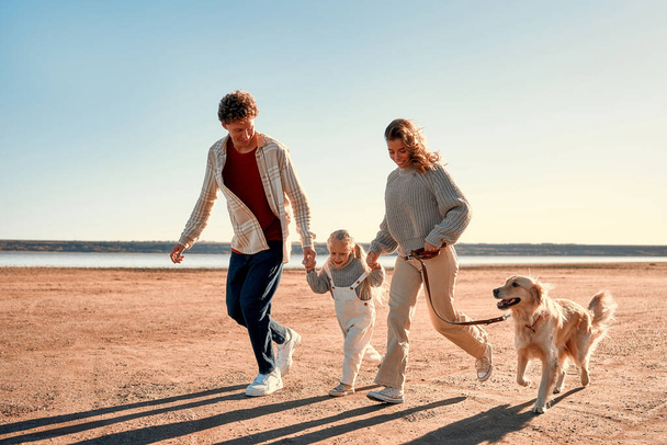 A young couple of parents with a little cute daughter and their beloved retriever dog walking and relaxing on a sandy beach in cool weather on their weekend. Family traveling with pet. - Photo, Image