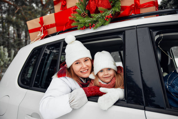 Vrolijk kerstfeest en gelukkig nieuwjaar. Gelukkig gezin met kinderen zittend in een auto geladen met dozen met geschenken en een kerstboom tegen de achtergrond van een bos met sneeuw weer. - Foto, afbeelding