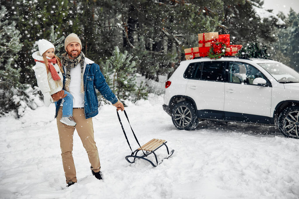 Mutlu Noeller ve yeni yıl. Mutlu çocuklu bir baba ve hediye dolu bir arabanın yanında duran bir kızak. Karlı bir havada bir ormanın arka planında bir Noel ağacı almaya gider.. - Fotoğraf, Görsel