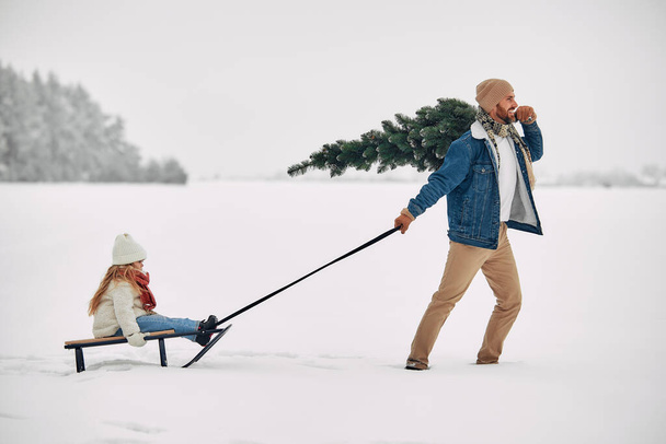 Καλά Χριστούγεννα και Καλή Χρονιά. Ένας ευτυχισμένος πατέρας κουβαλώντας την κόρη του σε ένα έλκηθρο και ένα χριστουγεννιάτικο δέντρο στον ώμο με φόντο το δάσος σε χιονισμένο καιρό, προετοιμάζεται για τις διακοπές. - Φωτογραφία, εικόνα