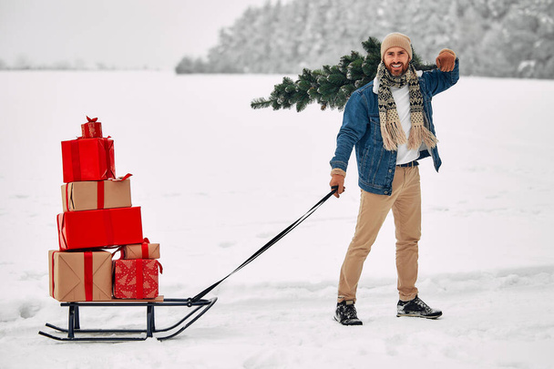 Merry Christmas and Happy New Year. A happy man is carrying a bunch of gift boxes on a sleigh and carrying a Christmas tree on his shoulder while walking along a snowy path. - Photo, Image