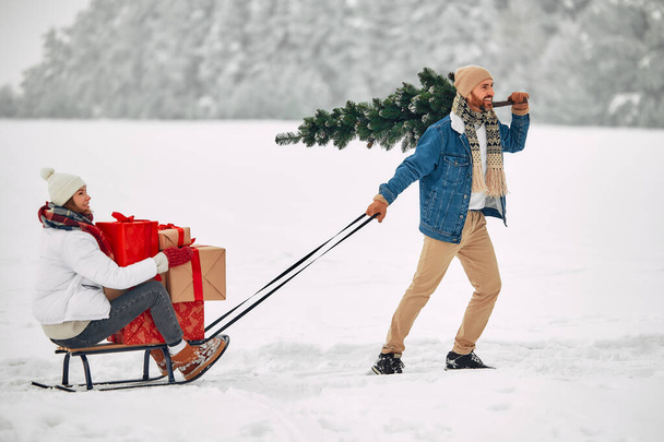 メリークリスマスとハッピーニューイヤー。 幸せな男はギフトボックスの束でガールフレンドをスリーに引っ張り,雪の道を歩いている間,彼の肩にクリスマスツリーを運びます. - 写真・画像