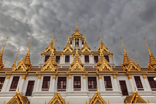 Templo Loha Prasat (Templo del Metal), en Bangkok, Tailandia. Edificio blanco rematado con adornadas agujas doradas. Cielo nuboso oscuro arriba.  - Foto, imagen