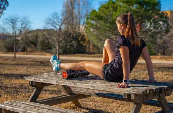 Fiatal, sportos futó lány, a hátán fekszik egy fa asztalon, a lábán gyakorolja az önmasszázst egy göröngyös fekete habos testmasszázsgörgővel, egy hegyi parkban napkeltekor.. - Fotó, kép