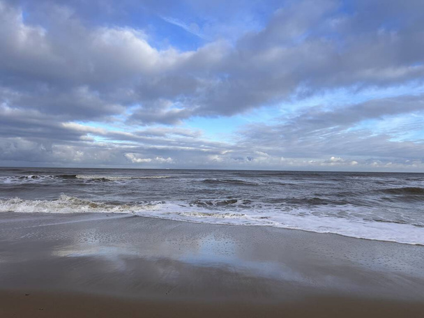 Beautiful landscape view of stunning wild sandy beach with white waves reflect clouds in ocean water against vast sea shore and grey dramatic blue skies with white grey clouds no people Winter day - Photo, Image