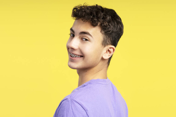 Nahaufnahme Porträt eines lächelnden attraktiven Jungen mit Zahnspange auf den Zähnen, der isoliert auf gelbem Hintergrund in die Kamera blickt. Gesundheitswesen, kieferorthopädisches Konzept - Foto, Bild