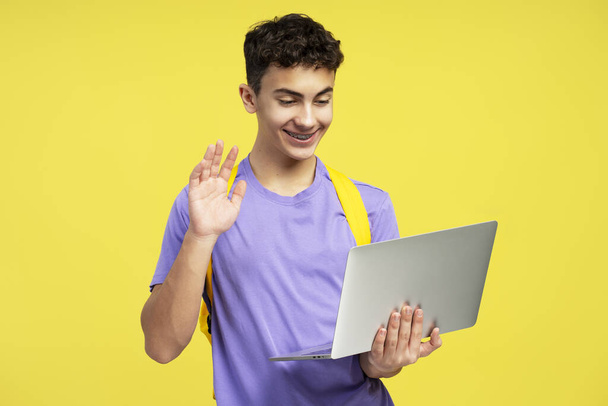Portré vonzó mosolygós tinédzser fiú visel elegáns alkalmi ruhák hátizsák, gazdaság laptop, integető kéz, amelynek video konferencia elszigetelt sárga háttérrel. Online oktatási koncepció - Fotó, kép
