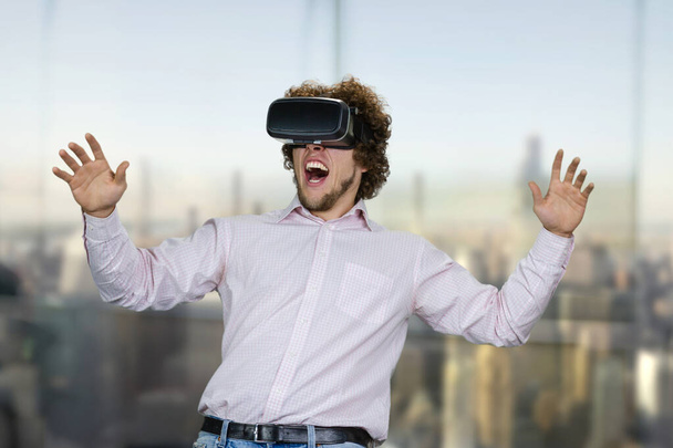 Retrato de jovem homem animado com cabelo encaracolado experimentando realidade virtual. Paisagem urbana turva no fundo. - Foto, Imagem