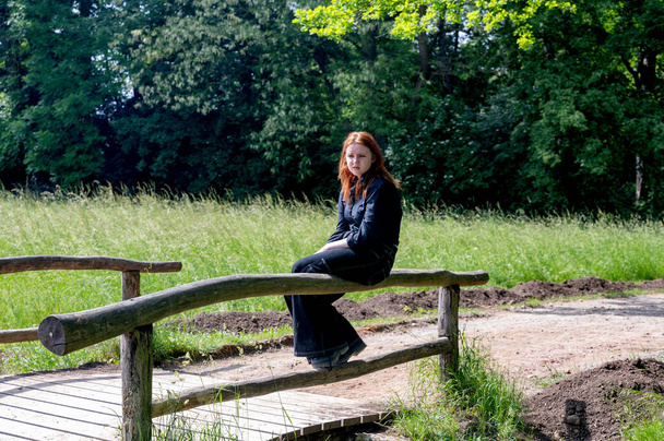Rudovlasá dospívající dívka smutně sedí na dřevěném zábradlí mostu v zelené přírodě za slunečného letního dne - Fotografie, Obrázek