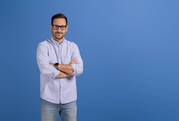 Άνδρας με αυτοπεποίθηση με σταυρωμένα τα χέρια και γυαλιά κοιτάζοντας την κάμερα σε μπλε φόντο - Φωτογραφία, εικόνα
