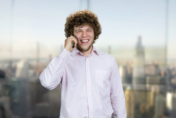 Beyaz gömlekli genç beyaz bir adamın portresi telefonda konuşuyor. Arka planda bulanık şehir manzarası olan kapalı pencere. - Fotoğraf, Görsel