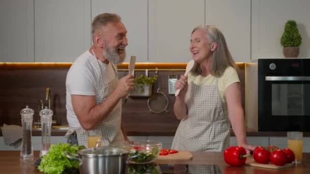 Hauska valkoihoinen pari rakastunut kypsä keski-ikäinen vanhempi vaimo ja aviomies eläkkeellä naimisissa perhe laulaa laulu lusikat keittiötarvikkeita iloinen naurava nainen mies onnellinen ruoanlaitto terveellistä ruokaa keittiössä - Materiaali, video