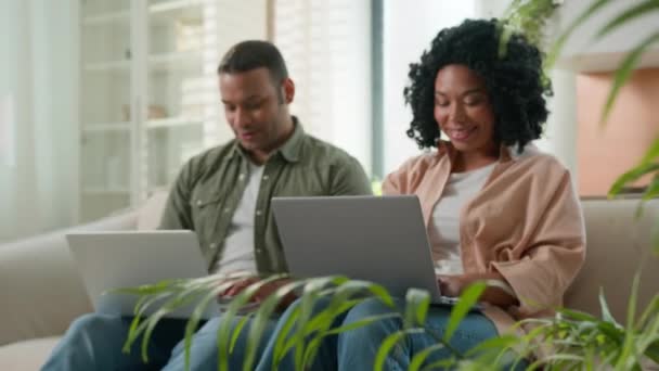 Мультирасовая пара влюбленных, работающих на ноутбуках дома афроамериканские семейные бизнесмены работают вместе на диване покупки онлайн на компьютерах глядя на камеру улыбается женщина и мужчина использовать устройства - Кадры, видео