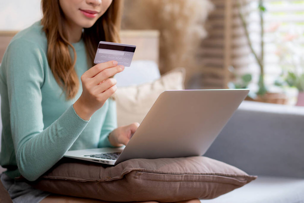 Młoda azjatycka kobieta siedzi na kanapie za pomocą laptopa zakupy online z karty kredytowej zakupu do Internetu, płatności kobiet z biznesu na kanapie, zakup i płatność, biznes i styl życia. - Zdjęcie, obraz