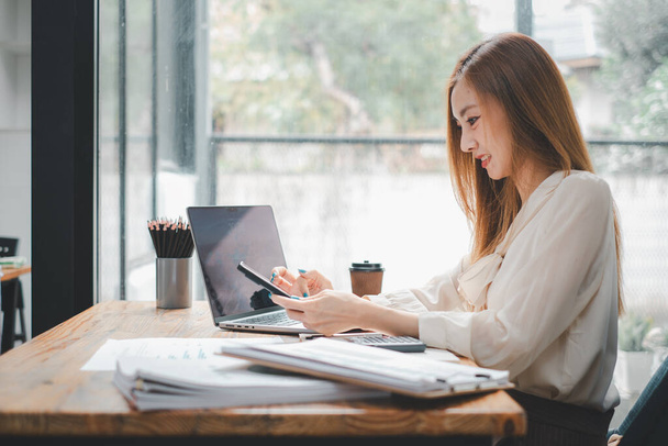 Привлекательная молодая женщина, работающая на мобильном телефоне и улыбающаяся, сидя на рабочем месте в офисе и глядя на ноутбук - Фото, изображение