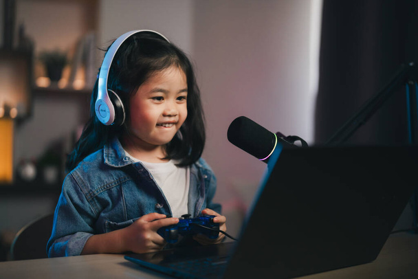 Ασιάτης influencer νεαρό κορίτσι χαμόγελο χαρούμενα χρησιμοποιώντας joystick παιχνίδι που παίζει live streaming χαιρετισμό βίντεο συνέδριο με φορητό υπολογιστή και φορώντας ακουστικά. Παιδιά streaming cast παιχνίδι σε απευθείας σύνδεση έννοια. - Φωτογραφία, εικόνα