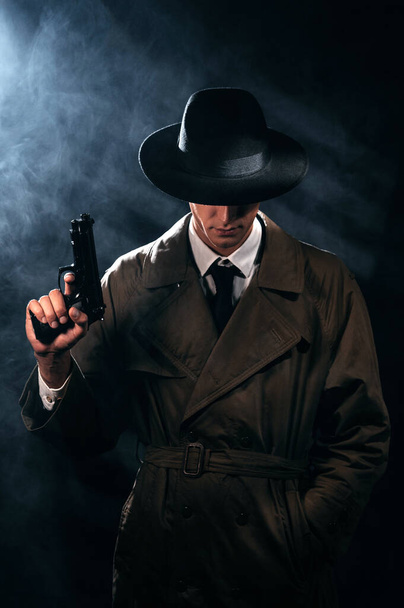 Silhouette of a male detective in a coat and hat with a gun in his hands. A dramatic noir portrait in the style of books and detective films of the 1950s - Foto, imagen
