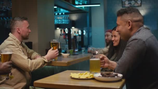 Imágenes medianas del grupo de amigos multirraciales sentados juntos en el pub celebrando y tintineando vasos de cerveza - Imágenes, Vídeo