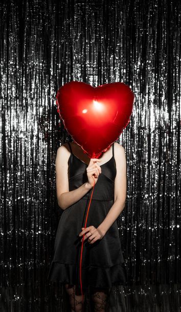 Stijlvolle jonge vrouw bedekt haar gezicht met een luchtballon in hartvorm op een donkere tinselachtergrond. Vrouw op Valentijnsdag. Symbool van de liefde. Kopieerruimte - Foto, afbeelding