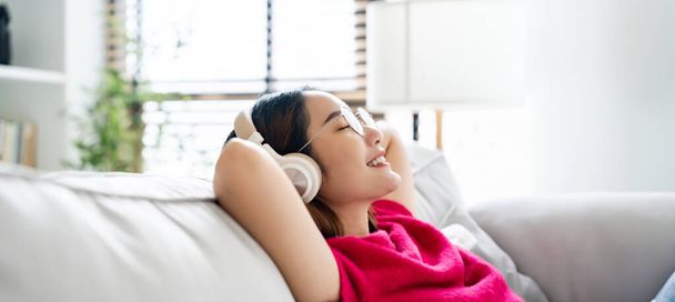 Relaxado jovem mulher asiática desfrutando de descanso no sofá confortável, calma atraente menina relaxante no sofá, respirar ar fresco com os olhos fechados, vestindo óculos e fone de ouvido, meditando em casa - Foto, Imagem